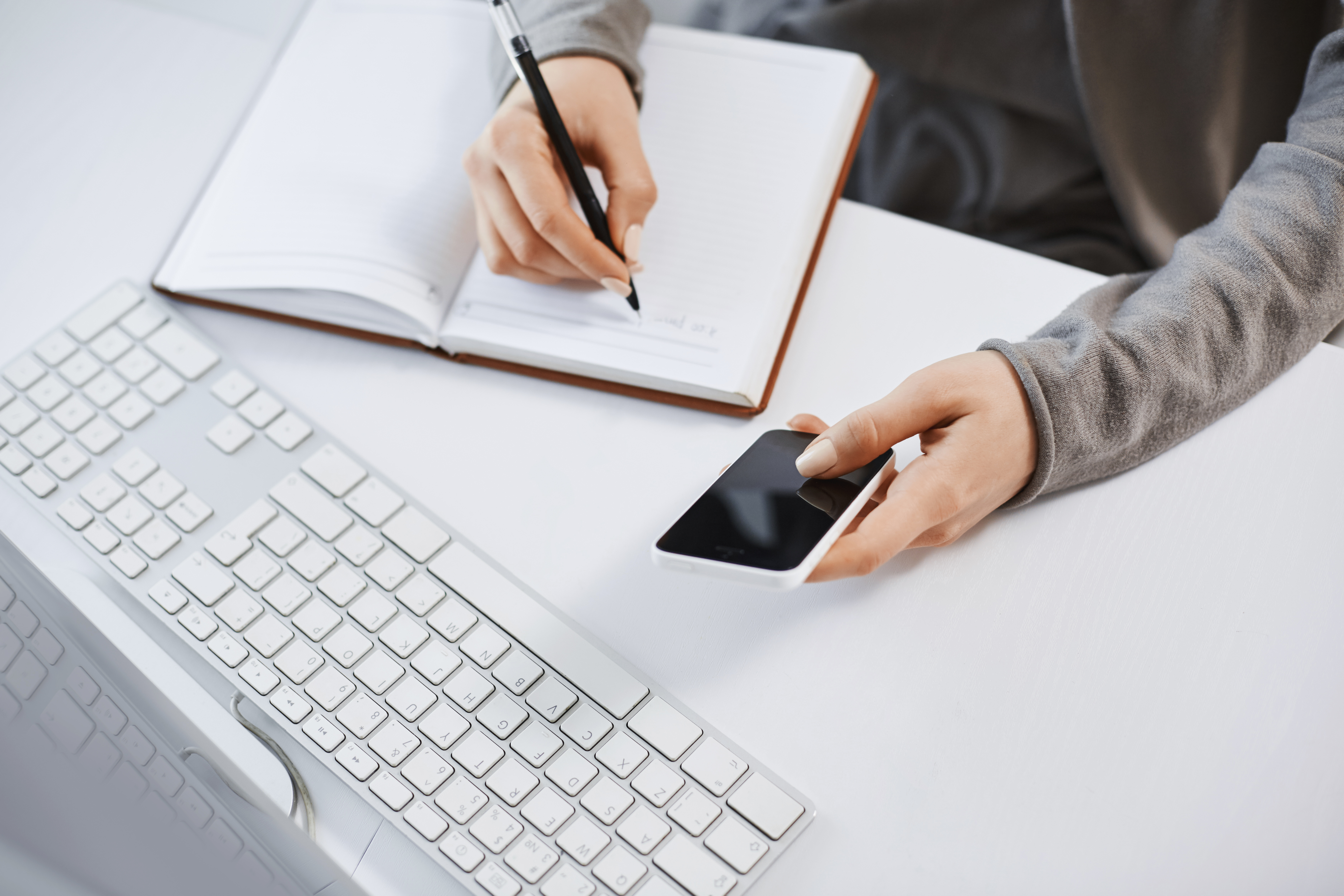 high-angle-shot-woman-hands-working-with-gadgets-cropped-shot-modern-female-holding-smartphone-while-writing-plan-notebook-sitting-near-keyboard-computer-having-tough-time-office